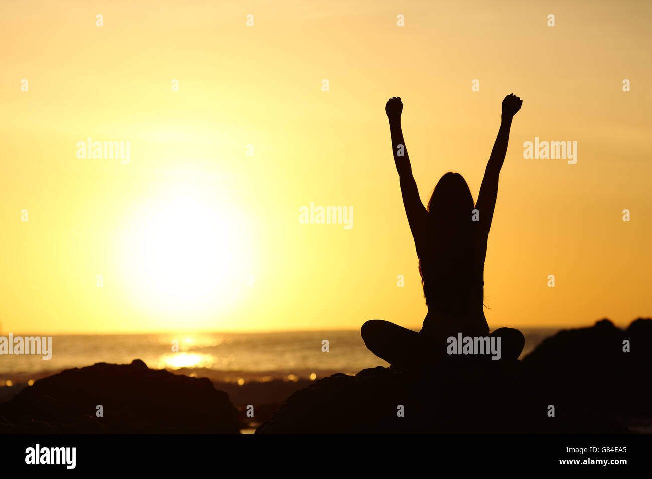 Vista posterior de un emocionado eufórica mujer silueta levantando los brazos y mirando al sol naranja en la playa al amanecer. Foto de stock