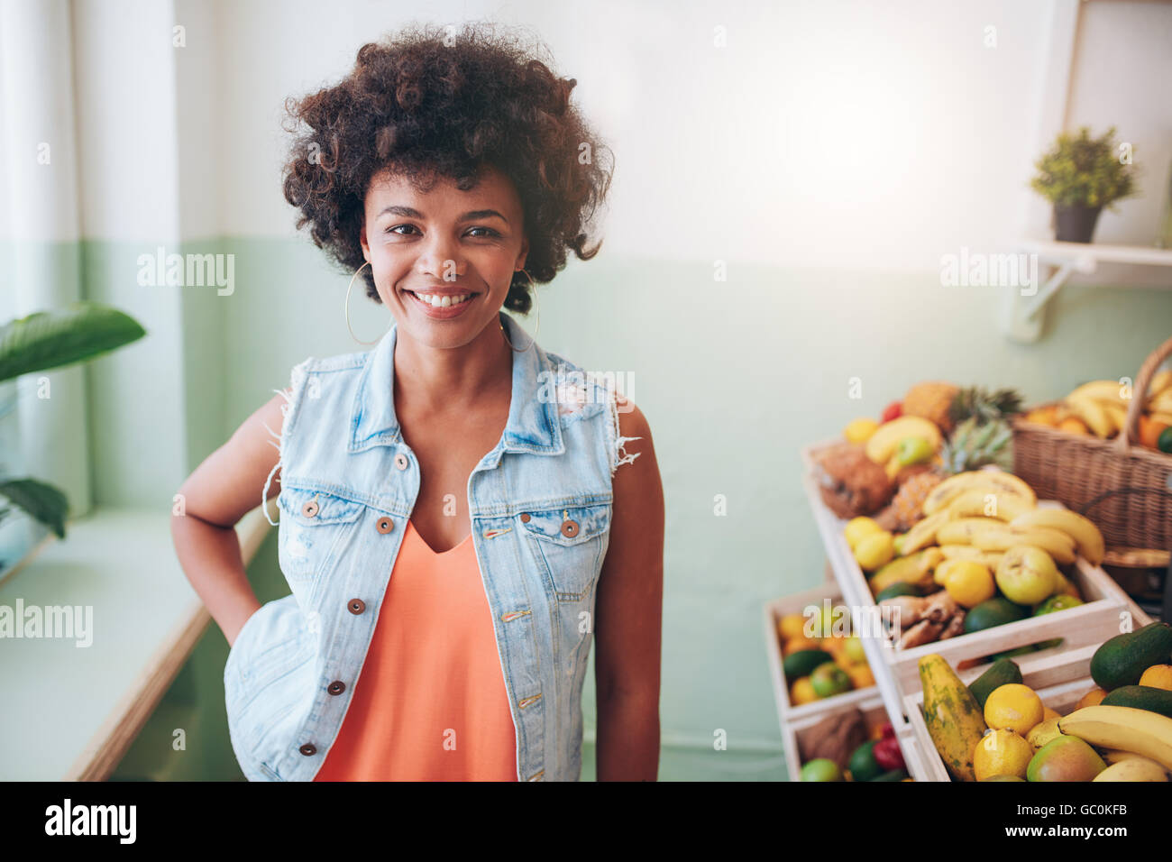 Retrato de joven feliz propietario juice bar mirando a la cámara y sonriendo. Modelo femenina africana en pie de frutas Estante en el cafe. Foto de stock