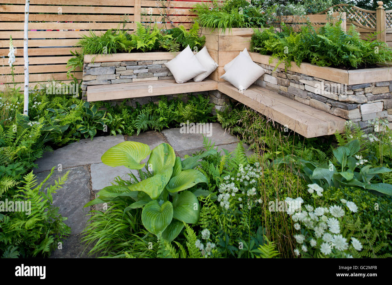 Una zona de asientos de piedra y madera rodeada de plantas en el interior de la ciudad jardín de gracia en Hampton Court Flower Show Foto de stock