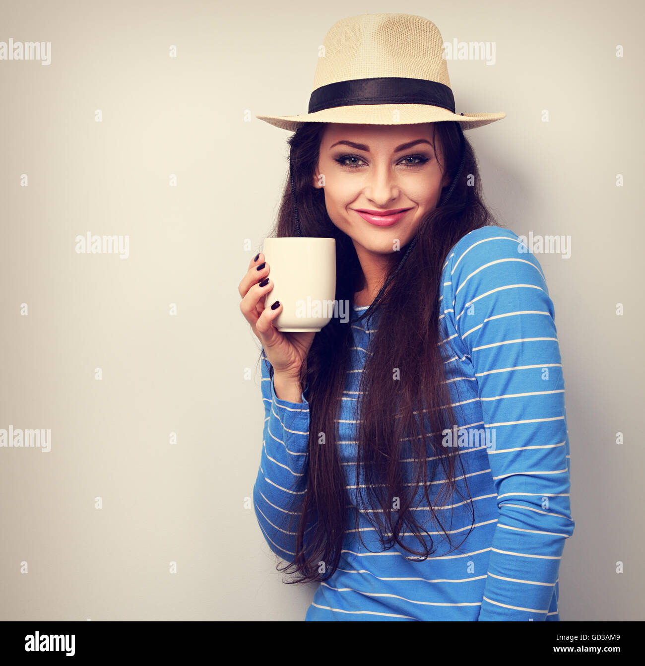 Lindo feliz mujer casual en el sombrero de paja sosteniendo en la mano una taza de té y sonriendo sobre fondo azul. Foto de stock