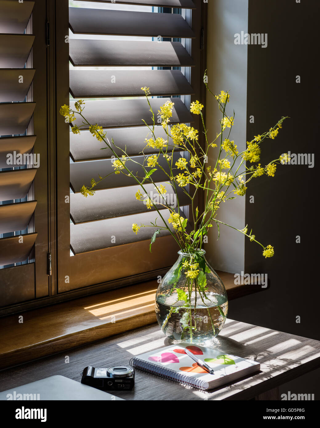 Detalle de jarrón con ramitas de flores amarillas con persianas de madera en segundo plano. Foto de stock
