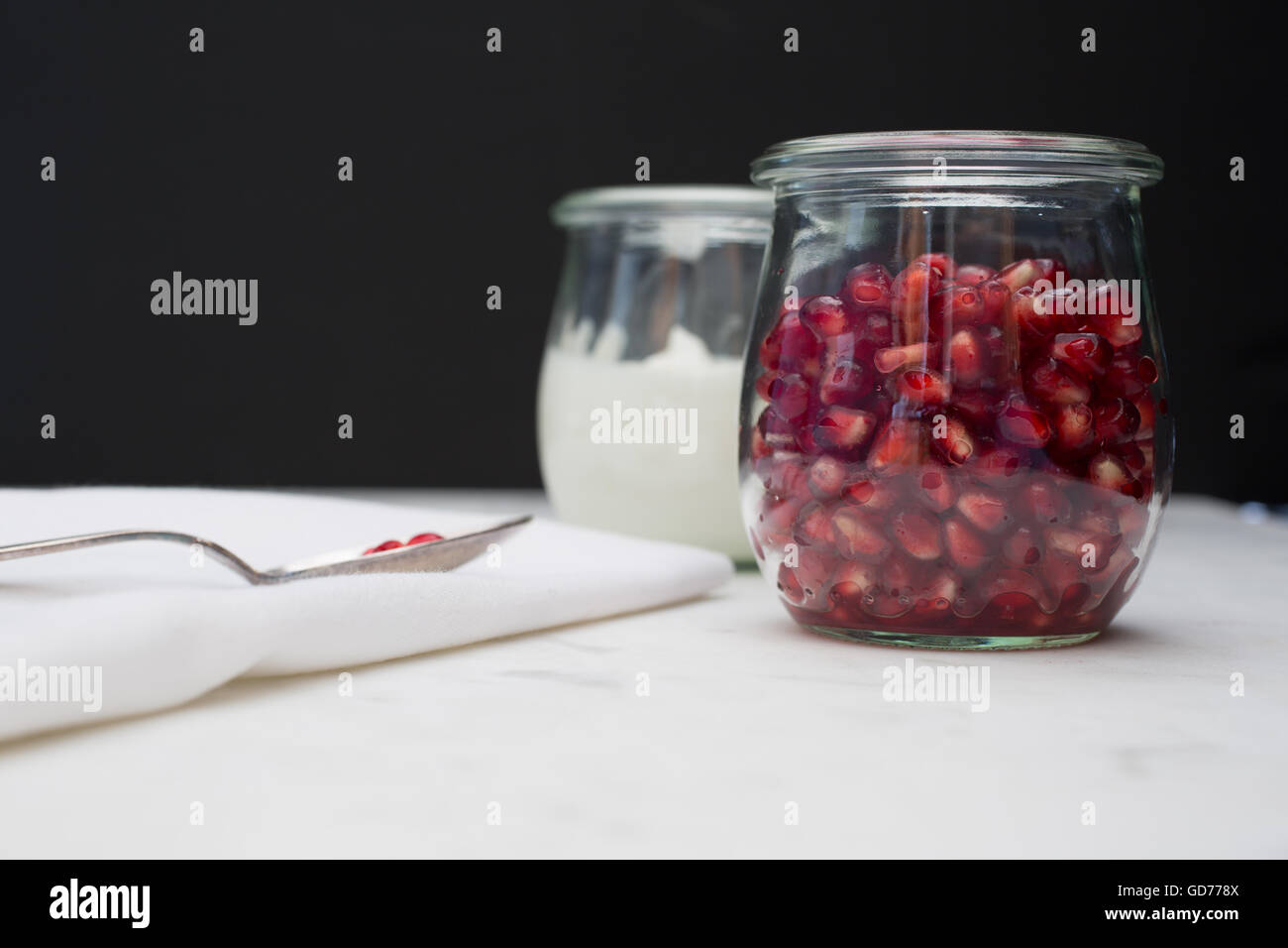 Vista frontal de un recipiente de vidrio de la granada arils (semillas), y el yogur. Unas pocas semillas descansan sobre una cuchara en una servilleta. Foto de stock