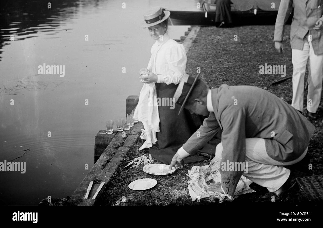 Picnic en el río Támesis...lavando platos y copas después. Una mujer joven y dos hombres con sombreros de paja. c1910. Fotografía por Tony Henshaw Foto de stock