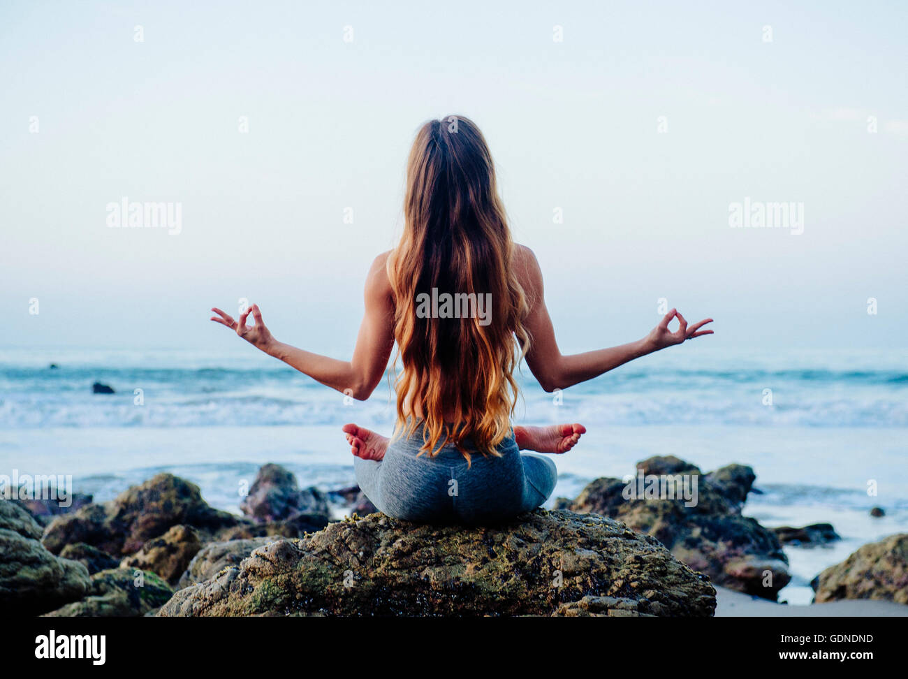 Vista trasera de la mujer joven con pelo largo practicando yoga loto plantean sobre rocas en la playa, Los Ángeles, California, Estados Unidos. Foto de stock