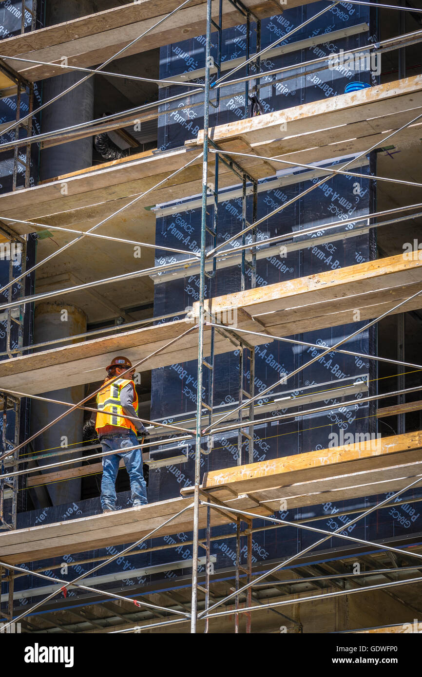Trabajador de la construcción en brillante chaleco de seguridad de pie sobre un andamio de alto edificio de oficinas en construcción Foto de stock