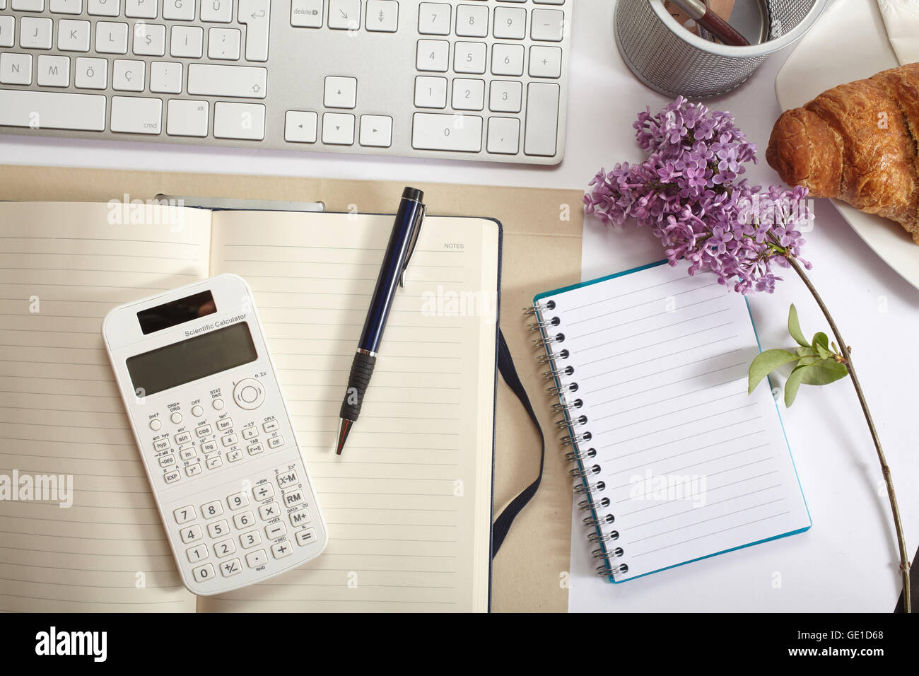 Vista superior de la oficina - croissant, teclado, calculadora, lápiz, flor y bloc de notas en el cuadro blanco Foto de stock