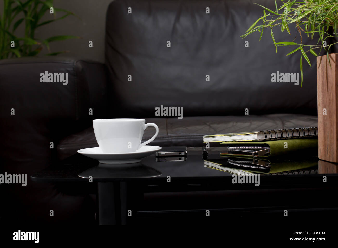 Taza de café blanco sobre una mesa de cristal de color marrón oscuro con un sofá en el salón Foto de stock
