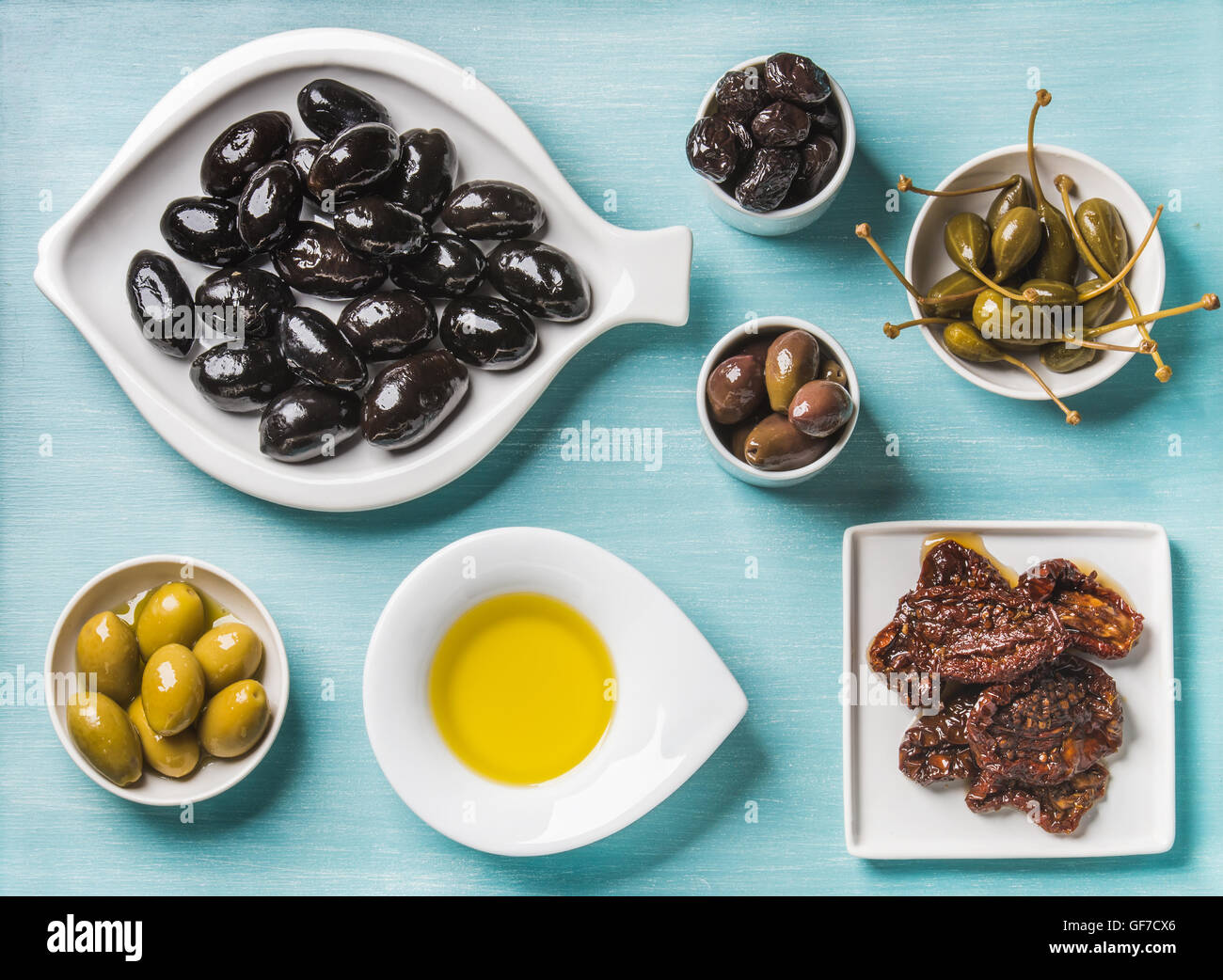 Surtido de aperitivos mediterráneos. Negro y verde griego de aceitunas, alcaparras, aceite de oliva, tomates secados al sol sobre fondo azul turquesa Foto de stock