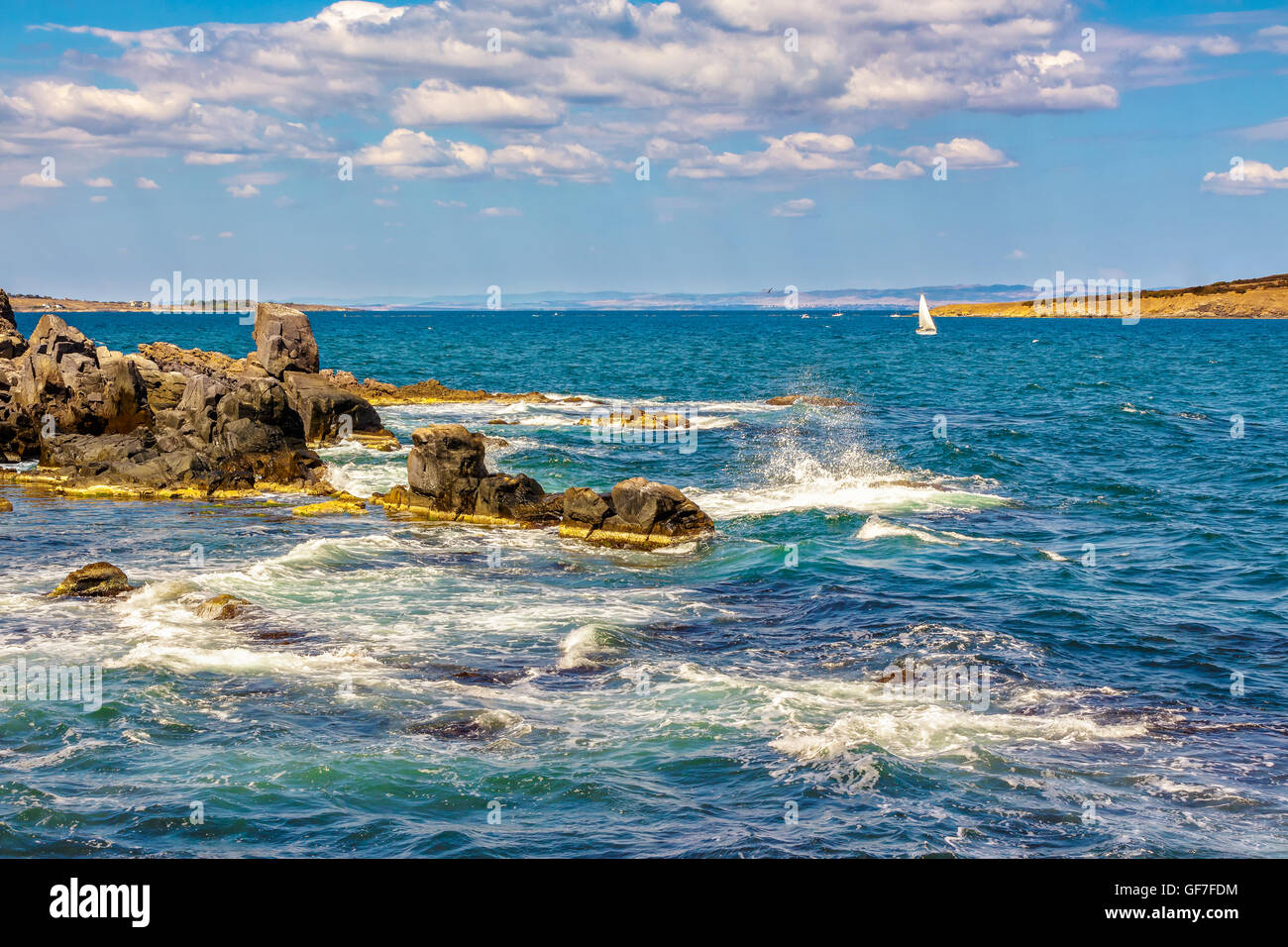 Pequeña embarcación navega a distancia en el mar. Las olas rompen en las enormes rocas en la orilla rocosa en el otro lado del Seascape Foto de stock