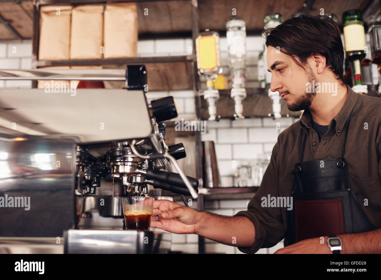 Vista lateral del varón barista prepara espresso en la cafetería. Joven en el delantal de hacer café con café en la cafetería. Foto de stock