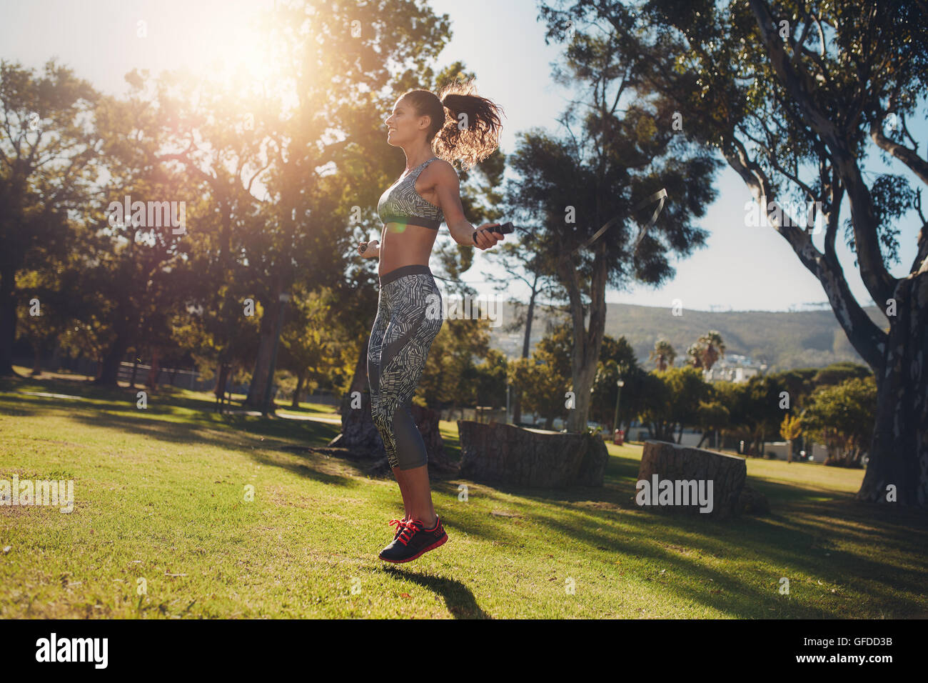Vista lateral del deportivo mujer saltando en un parque en una mañana soleada. Colocar mujeres atléticas y hacer ejercicio con una soga para saltar sobre las gra Foto de stock