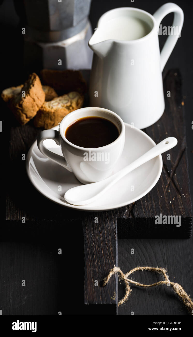 Juego de café italiano para el desayuno. Taza de café expreso caliente,  creamer con leche y moka cantucci olla sobre tablero de madera rústica  oscuro sobre fondo negro Fotografía de stock -