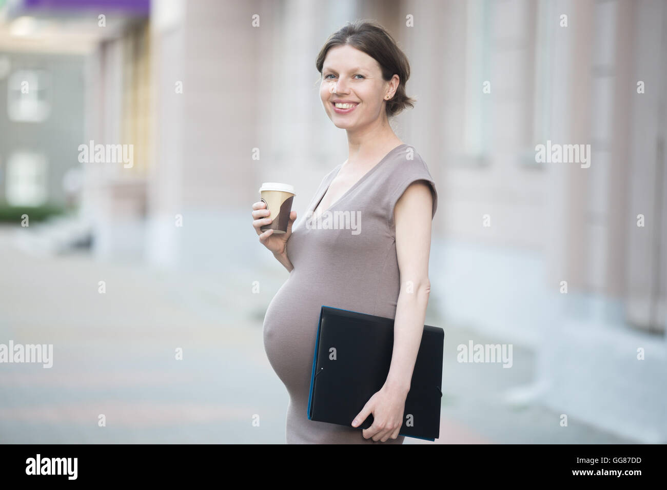 Mamá activa. Retrato de feliz empresa joven mujer embarazada mirando a la cámara mientras está de pie en la calle con la carpeta de documentos Foto de stock