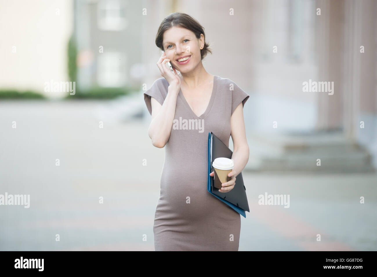 Active el embarazo. Retrato de mujer de negocios jóvenes embarazadas felices caminando en la ciudad con la carpeta Documentos y hacer llamada Foto de stock