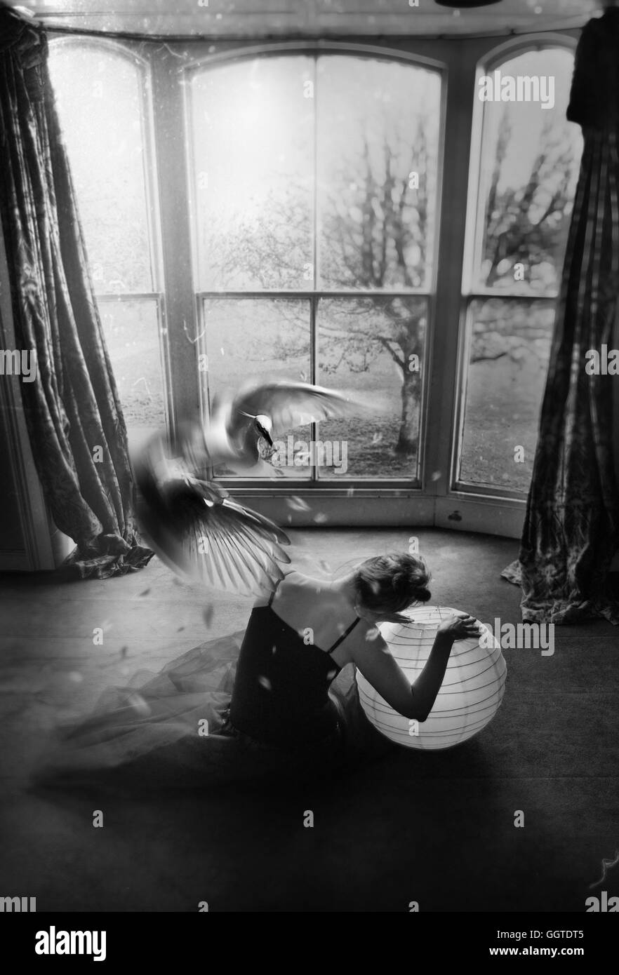 Vista posterior de la joven sentada en el suelo sujetando una bola con un gran pájaro blanco volando Foto de stock