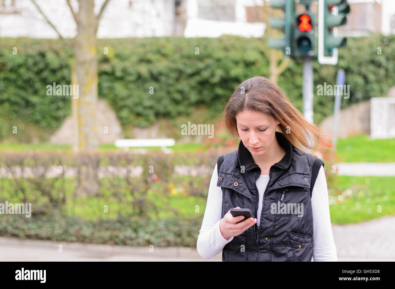 Mujer joven comprobación de mensajes de texto en su teléfono móvil mientras camina a lo largo de la calle en un paso de peatones en rojo Foto de stock