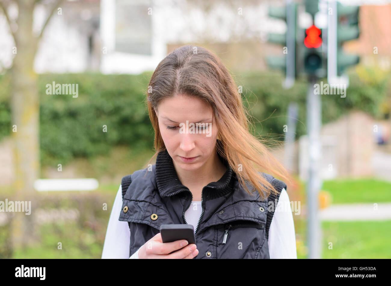 Mujer joven comprobación de mensajes de texto en su teléfono móvil mientras camina a lo largo de la calle en un paso de peatones en rojo Foto de stock