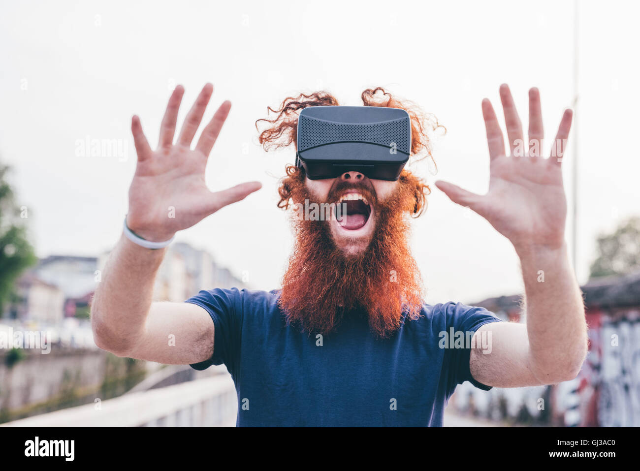 Retrato de macho joven hipster con pelo y barba roja gritando mientras llevaba casco de realidad virtual Foto de stock