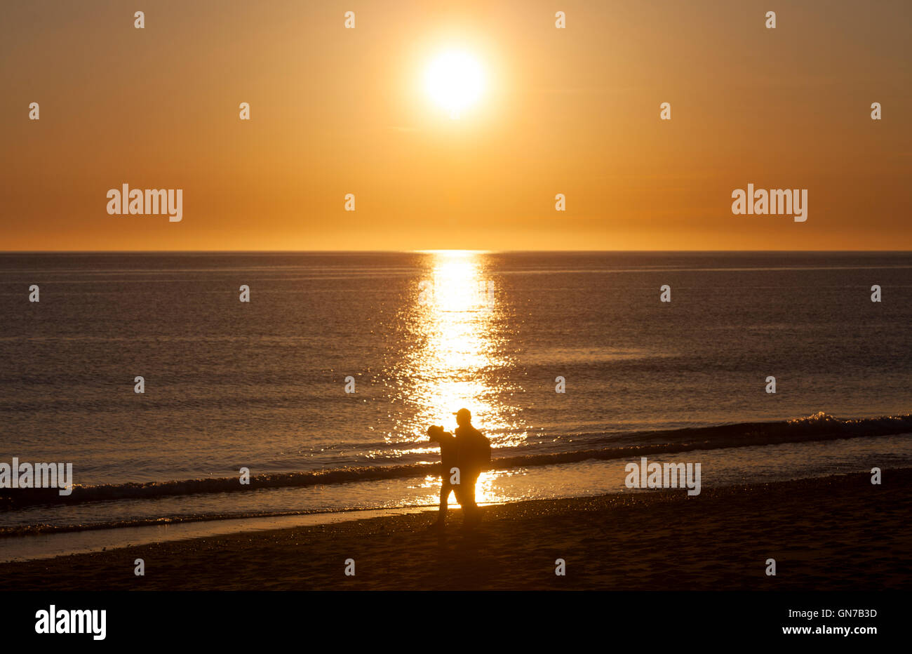 Una puesta de sol sobre el océano y la silueta de 2 personas caminando por la playa. Foto de stock