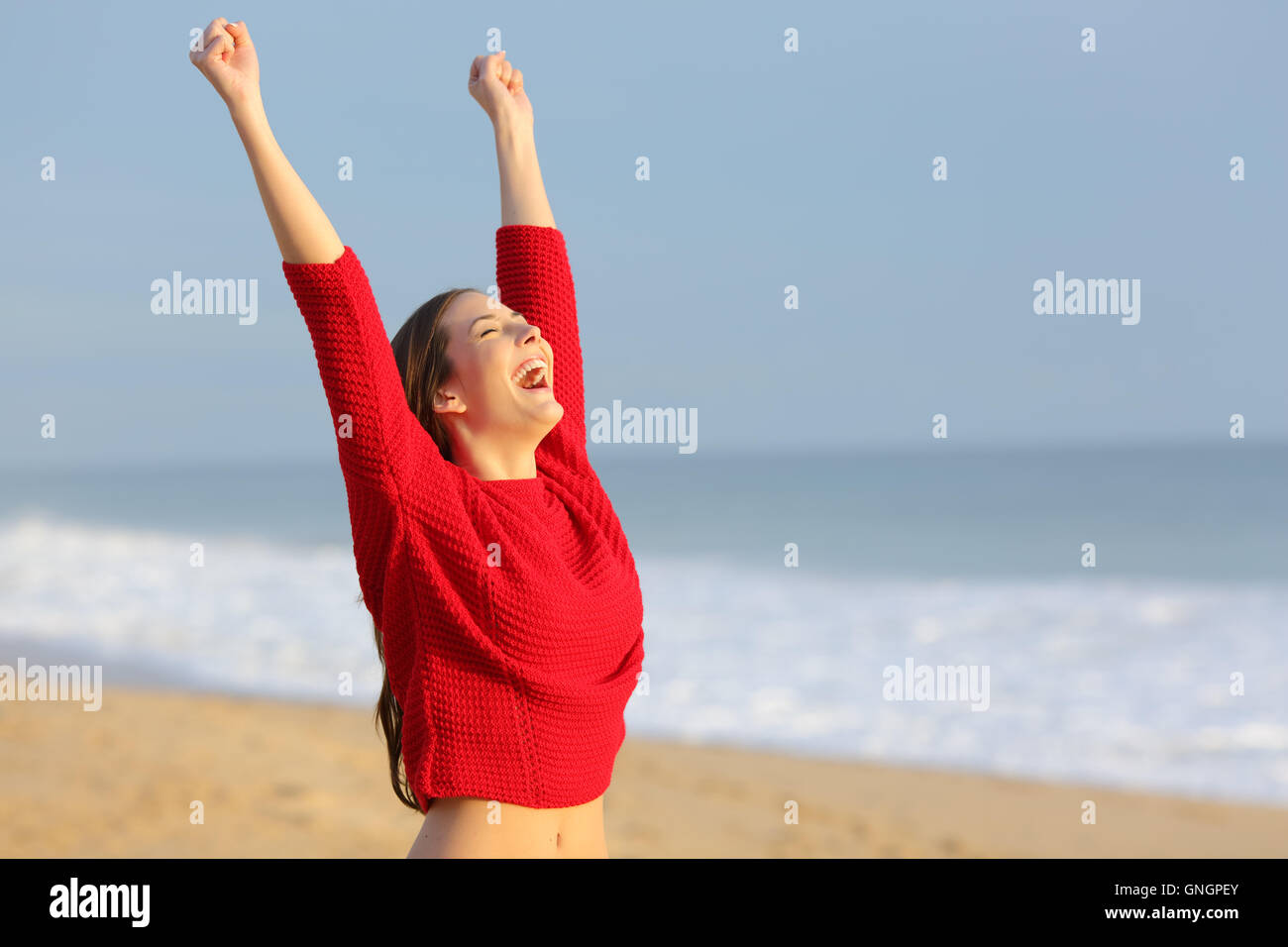 Feliz gracioso Emocionados mujer vistiendo un suéter color rojo levantando brazos eufóricos por la playa al atardecer con una luz cálida Foto de stock