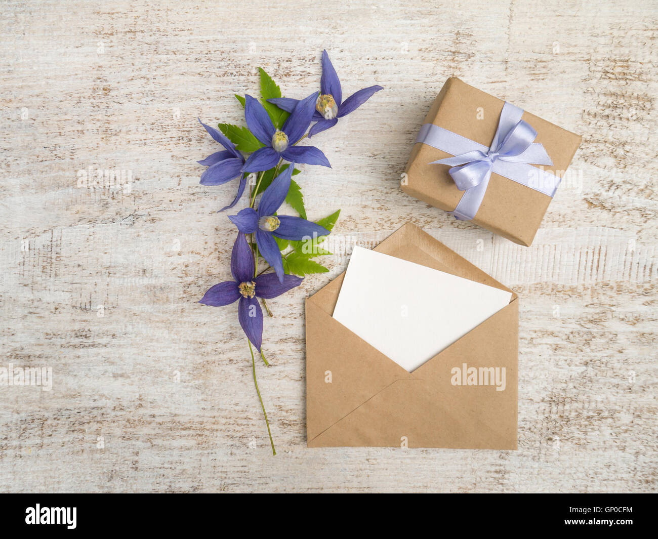 Caja de regalo envuelto en papel kraft marrón con lazo de raso violeta, flores azules y sobres con tarjeta de felicitación en la madera pintada Foto de stock