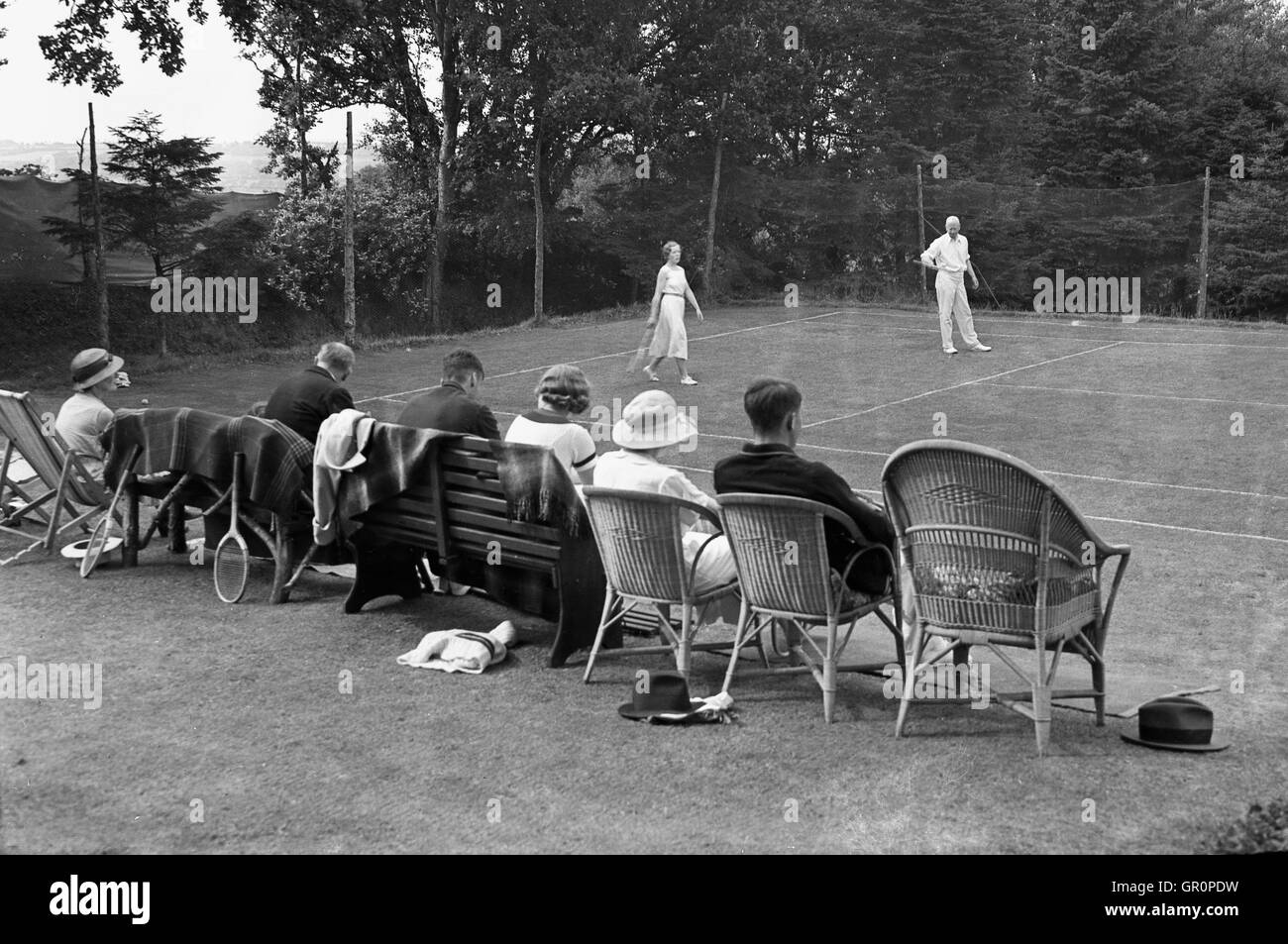 1930, históricos, par jugar en un fuera de pista de tenis de hierba en Spreyton village, Devon, Inglaterra, observados por un pequeño número de espectadores sentados. Foto de stock