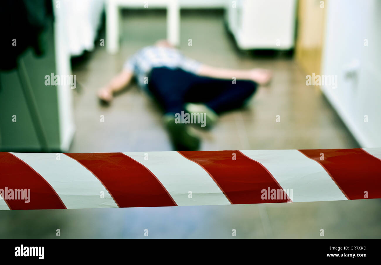Detalle de una escena de crimen con el cadáver de un joven acostado en el piso en el interior, con una cinta roja y blanca de la policía en el fo Foto de stock
