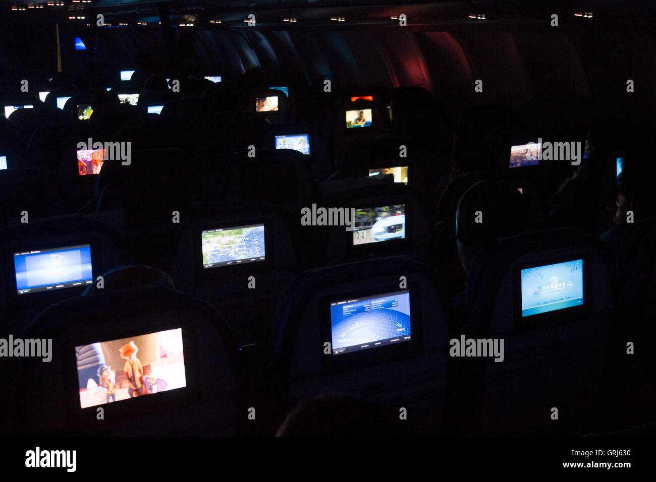 Interior de la cabina de una aeronave con luces atenuadas durante la noche vuelo nocturno con pantallas de TV personales sobre respaldos de los asientos Foto de stock