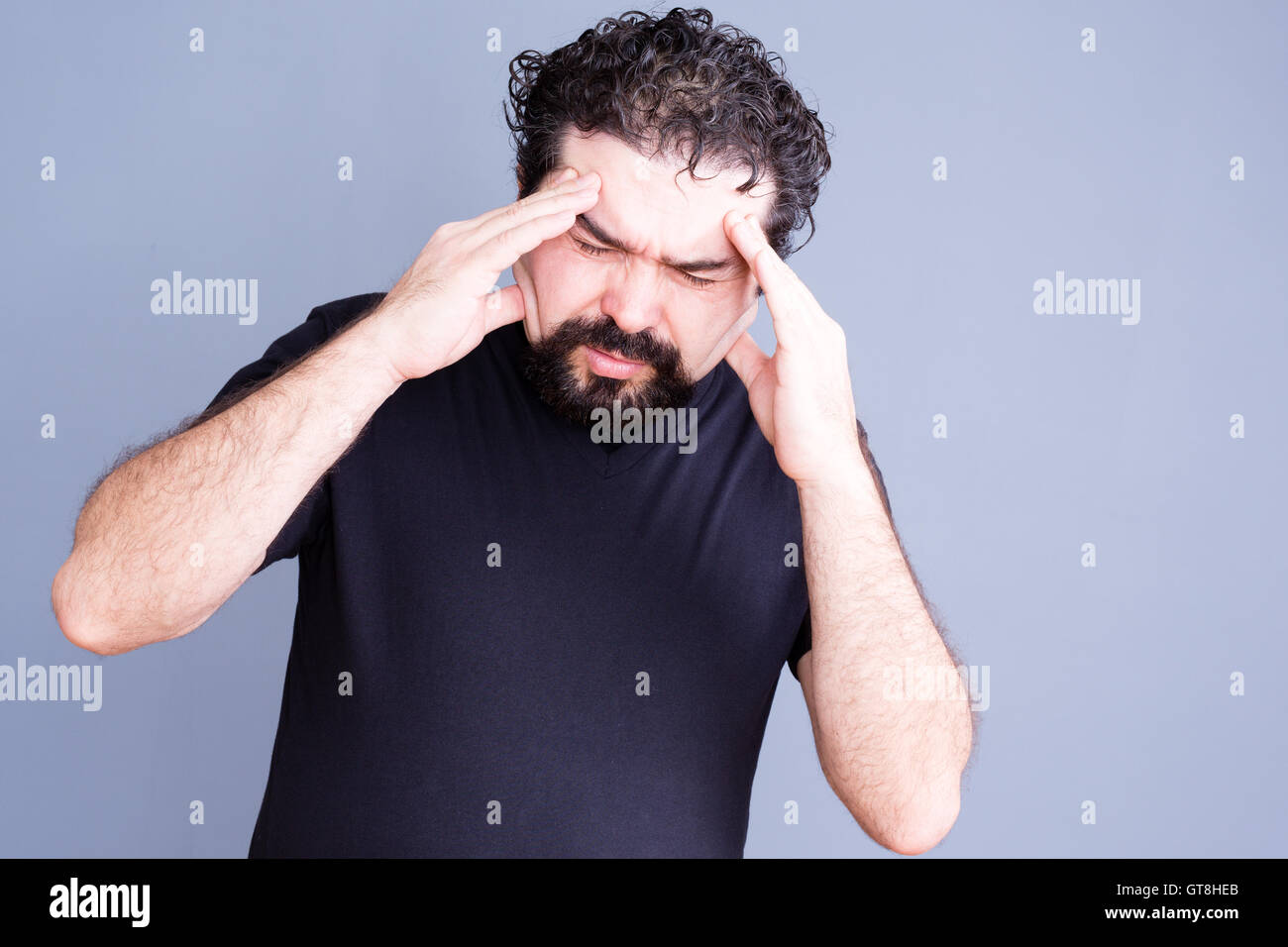 Sobrecargado de trabajo único hombre vestido con barba frota su frente en dolor como si sufre de dolor de cabeza o estrés sobre fondo gris Foto de stock