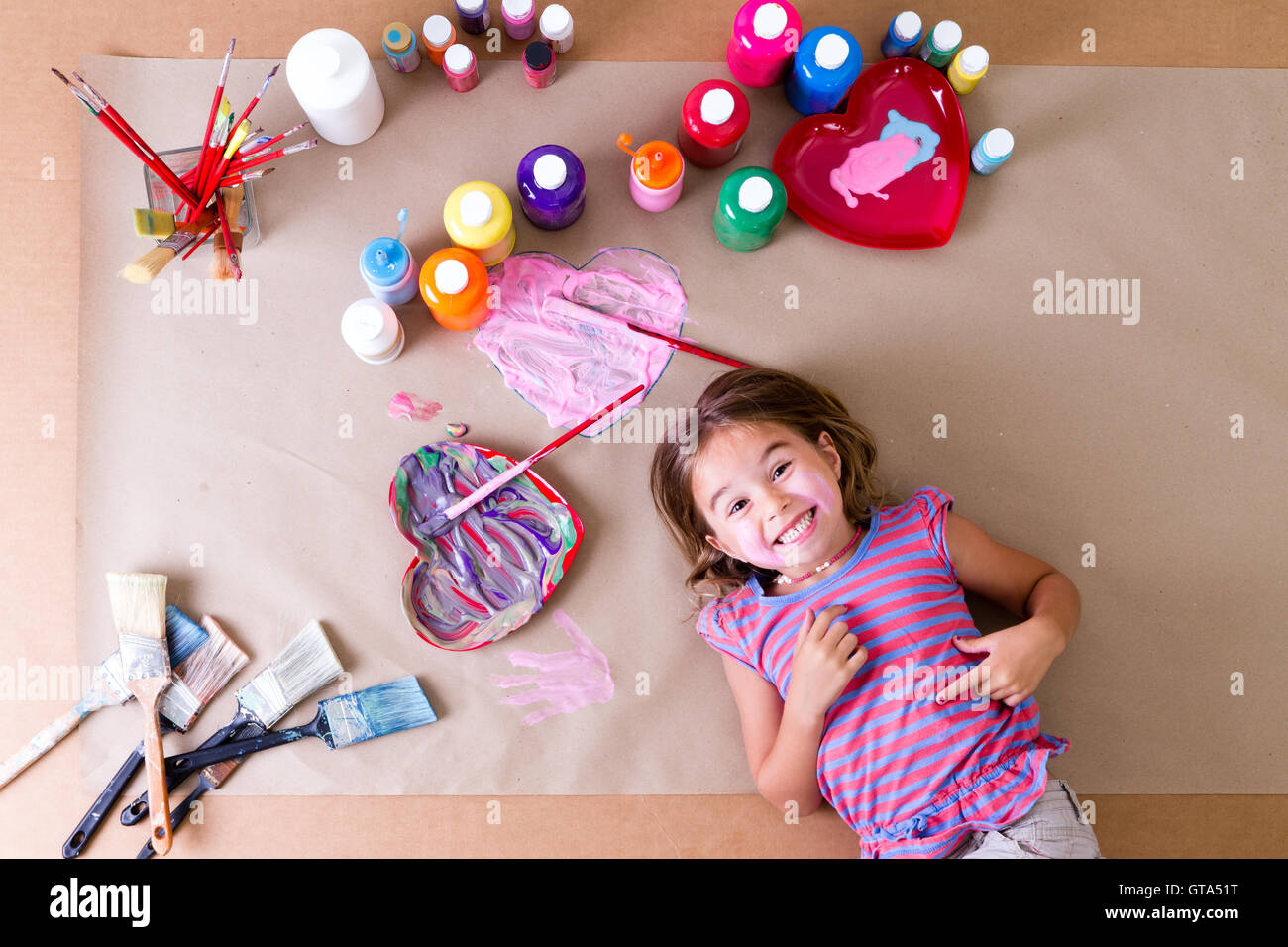 Feliz pícara niña artista con su arte suministros tumbado rodeado de coloridos botes de pintura, pinceles y bastante corazón desig Foto de stock