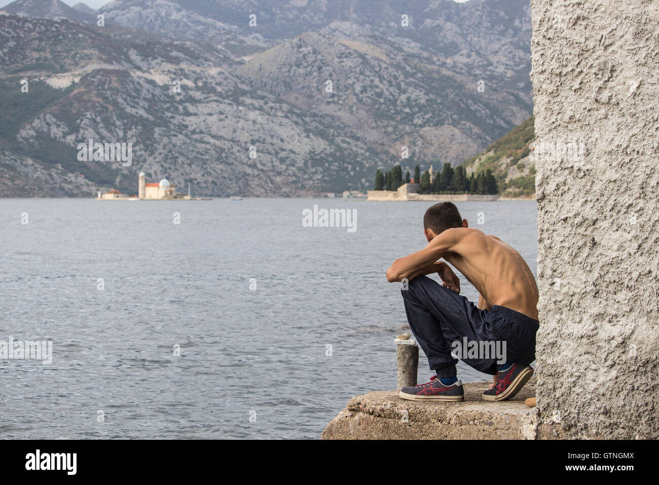 La bahía de Kotor, Montenegro - Skinny hombre agachado hacia abajo a la orilla del mar observa el islote virgen de las rocas (Gospa od Skrpjela) Foto de stock