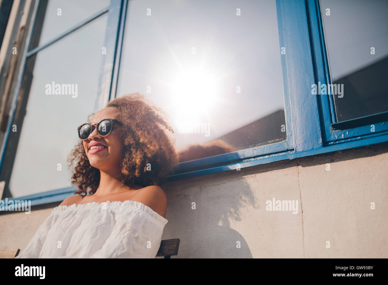 Disparó al aire libre de feliz joven femenina africana sentados al aire libre y sonriente. Mujer con gafas de sol sentado fuera de su casa. Foto de stock