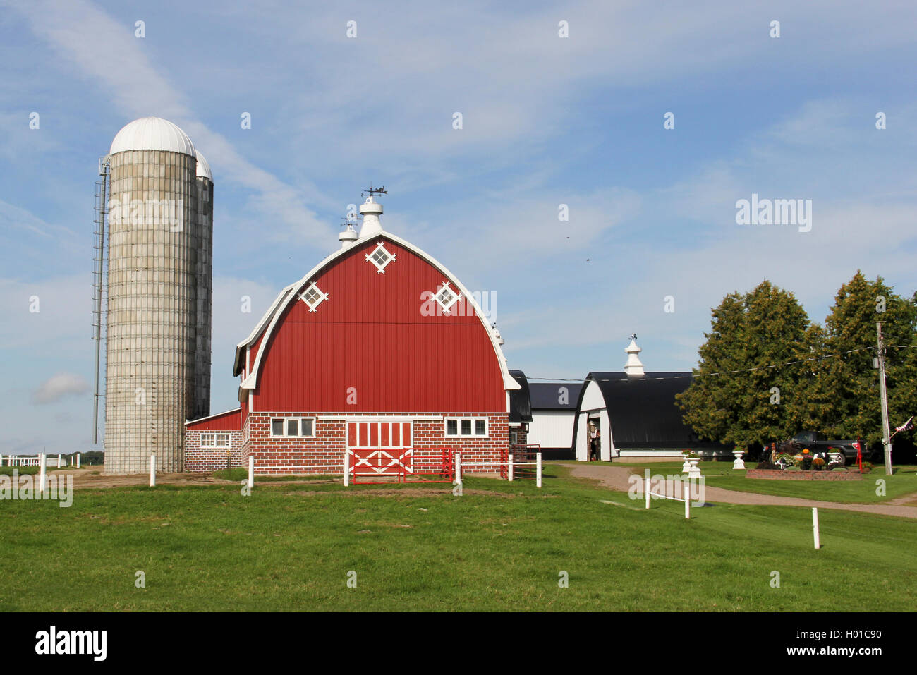 Granero rojo con silo y una cerca blanca con el pasto verde y árboles Foto de stock