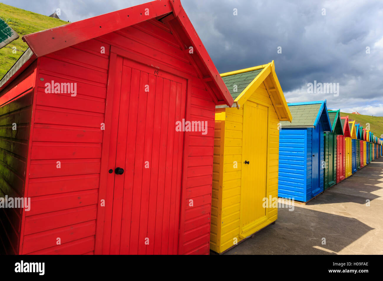 Fila de coloridas casetas de playa y sus sombras, con acantilados de hierba, West Cliff Beach, Whitby, North Yorkshire, Inglaterra, Reino Unido. Foto de stock