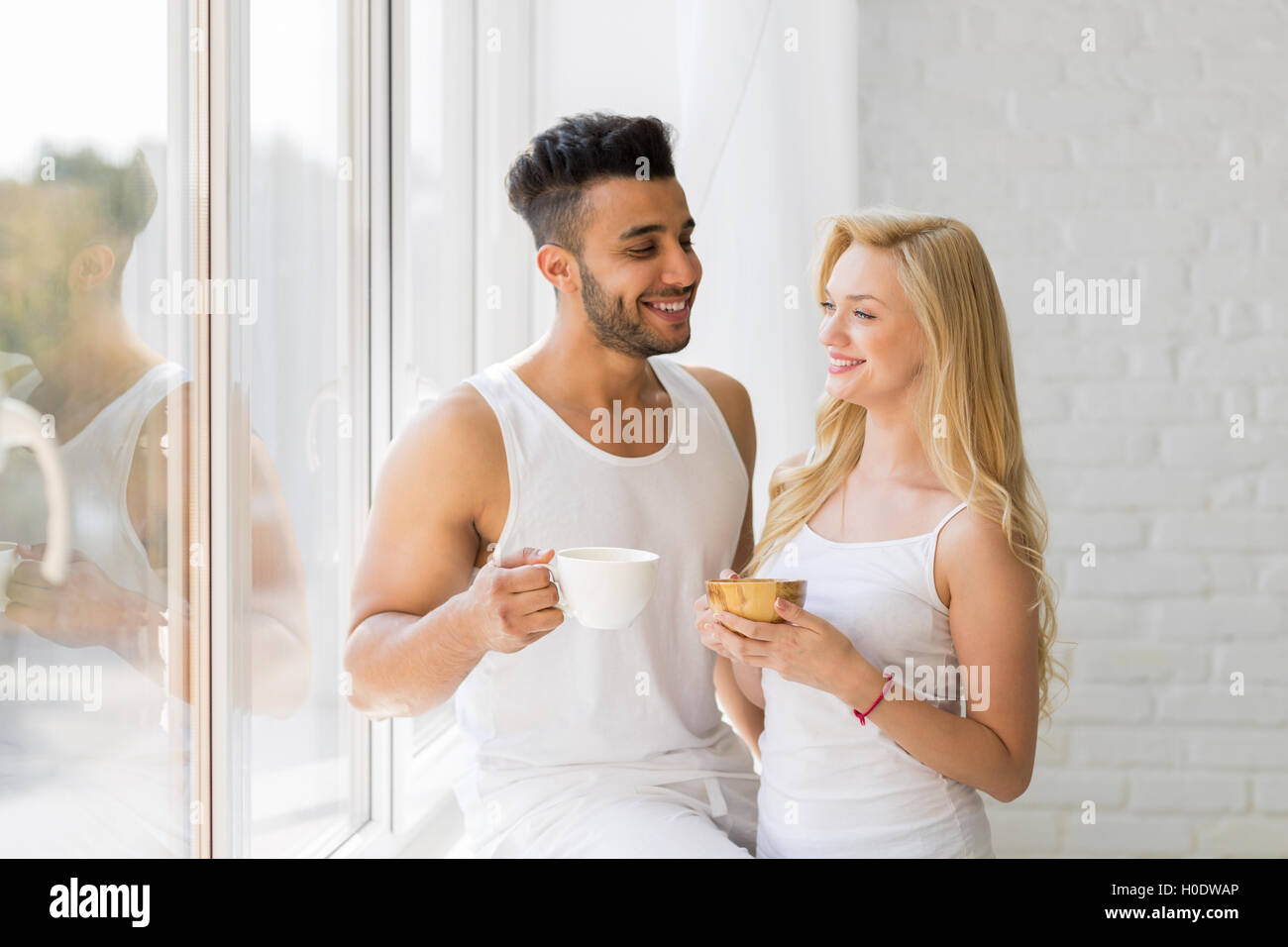 Hermosa joven pareja permanezca cerca de ventana grande, beber de la taza de café por la mañana, sonrisa feliz al hombre y a la mujer hispana Foto de stock