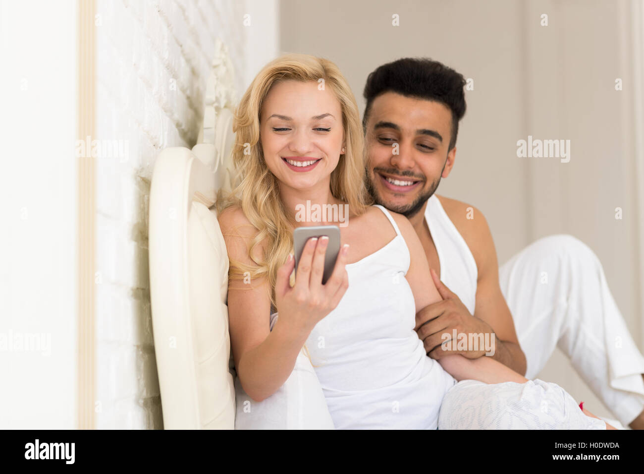 Pareja joven acostado en la cama, sonrisa feliz al hombre y a la mujer hispana utilizando la celda teléfono inteligente. Foto de stock