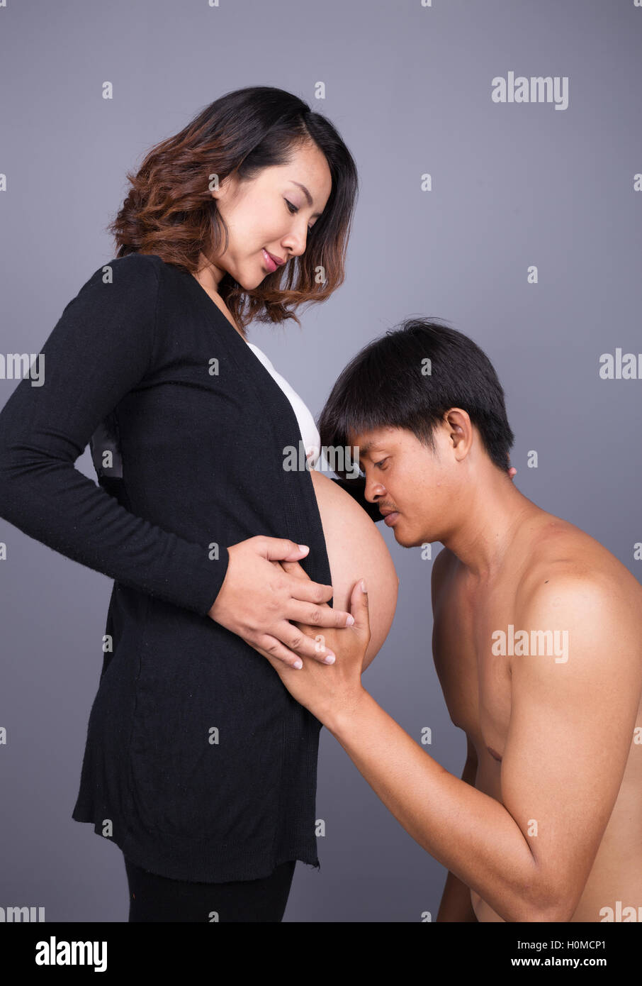 Joven besando a su esposa embarazada del vientre gris sobre fondo de pared Foto de stock