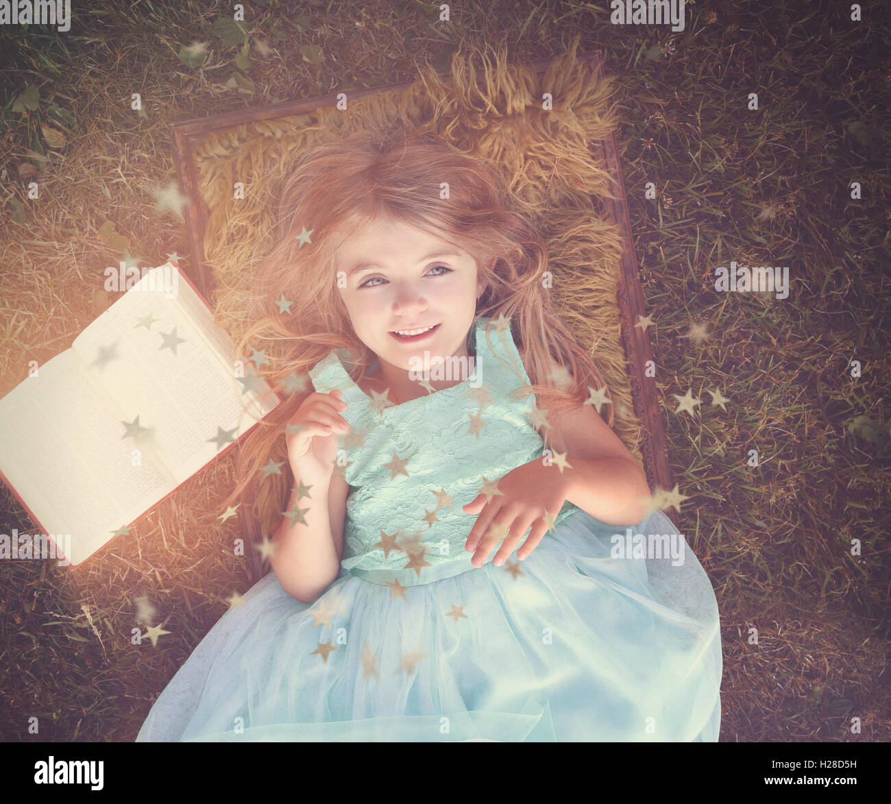 Una bonita niña feliz está sentando en el césped fuera con un libro de cuentos para un misterio, imaginación o concepto de cuento de hadas. Foto de stock