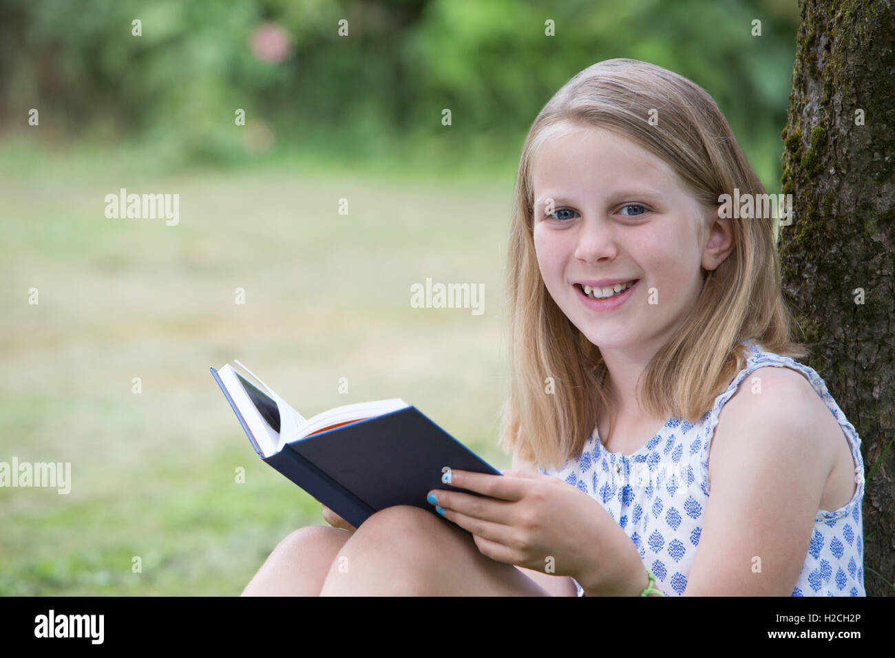 Chica sentada contra el árbol en el jardín del libro de lectura Foto de stock