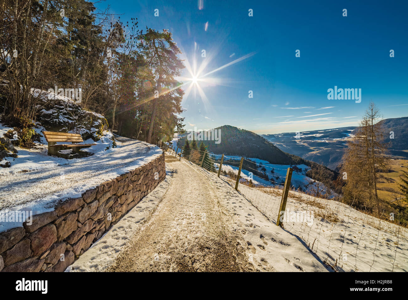 Bench en carretera de montaña con vistas de los tejados cubiertos de nieve de una aldea alpina en Italia Foto de stock