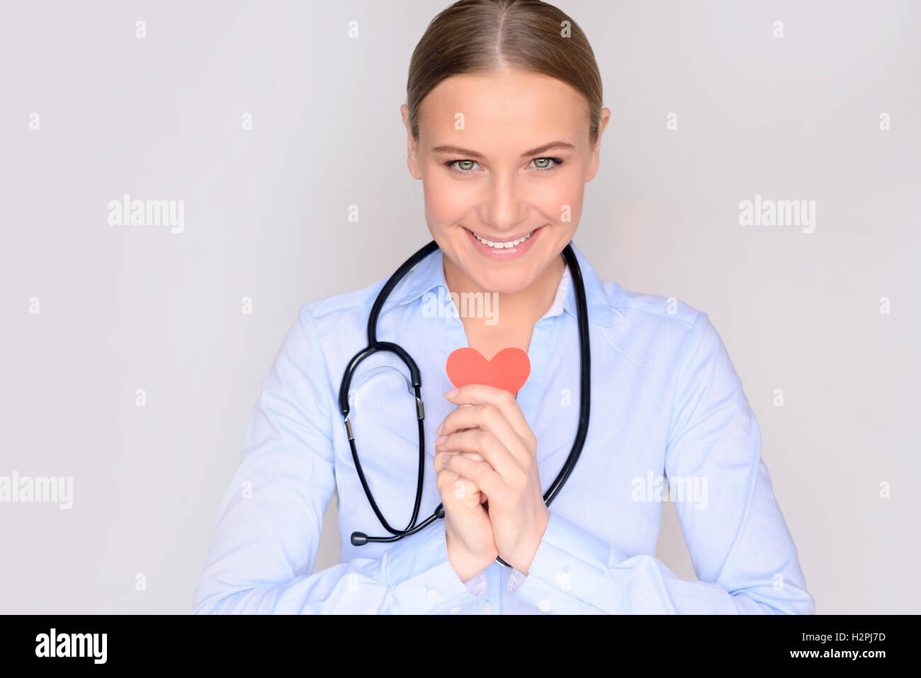 Retrato de una mujer sonriente lindo cardiólogo holding en manos poco papel de corazón y vistiendo bata médica y estetoscopio Foto de stock
