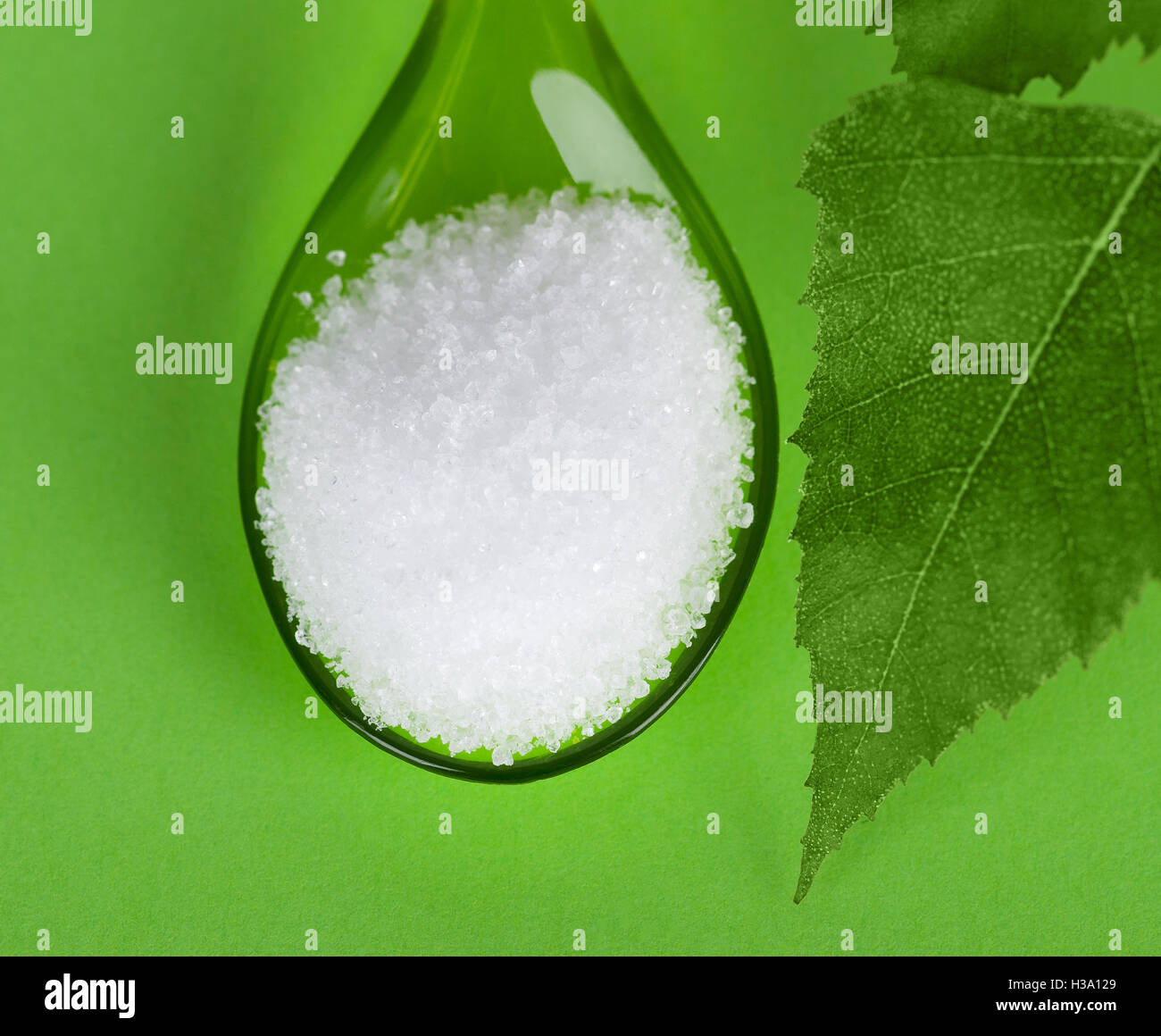 El Xilitol azúcar de abedul en cuchara de plástico con hojas de abedul sobre fondo verde. Sustituto de alcohol de azúcar blanco granulado. Foto de stock
