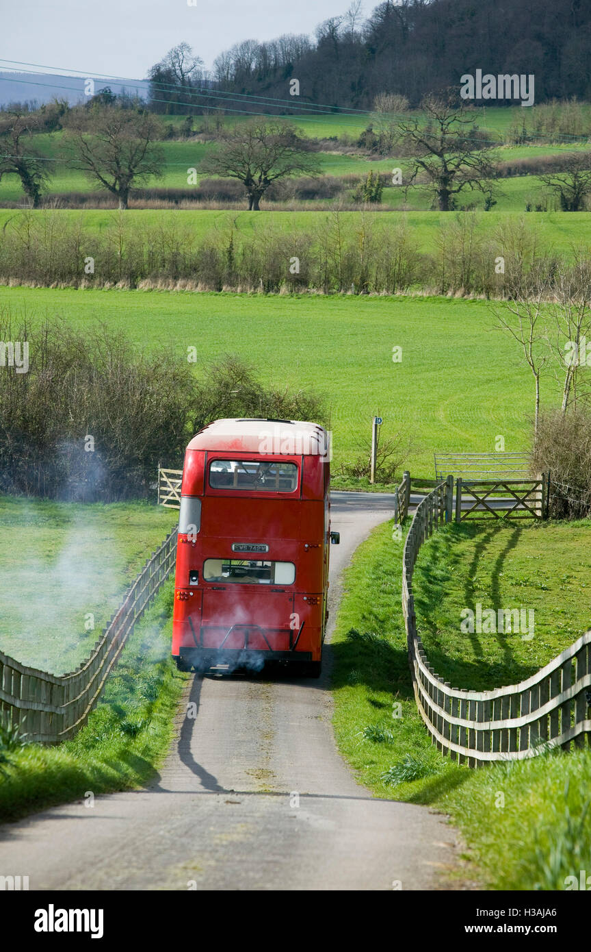 Un viejo double decker bus rojos de Londres Biritish conduciendo a través de la campiña en un viaje tomando pasado colinas,fieldds adn árboles. En una escena remeniscent de la famosa película de vacaciones de verano con Cliff Richard el bus puede sacar a la gente de vacaciones o de vacaciones. Foto de stock