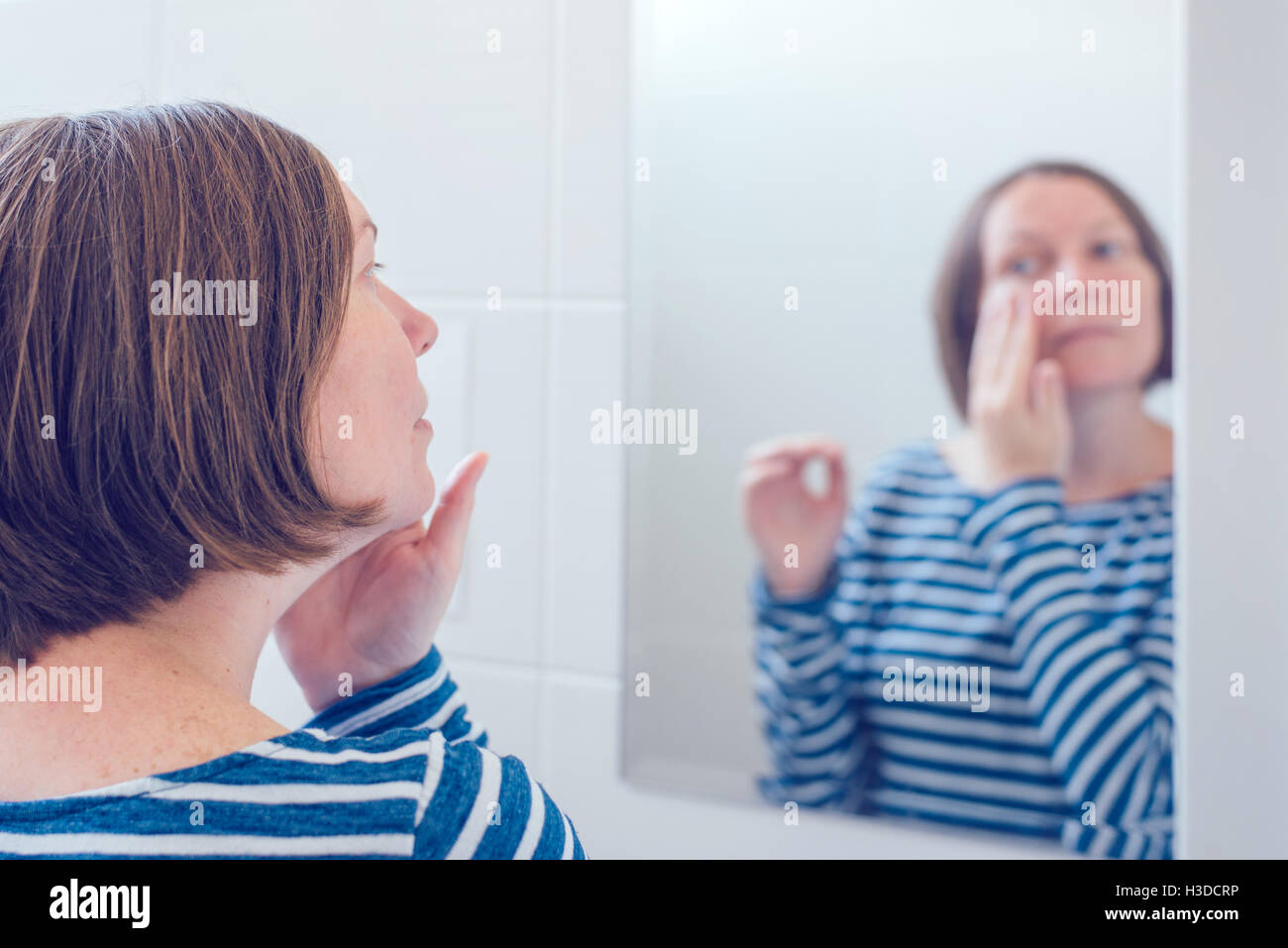 Mujer adulta joven aplicar crema anti-envejecimiento delante del espejo del baño, el enfoque selectivo Foto de stock