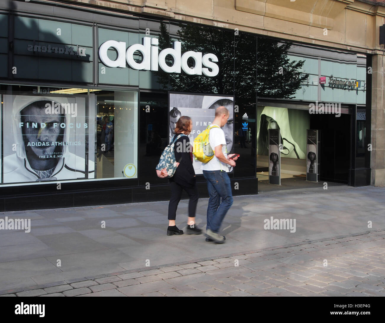 Tienda de ropa deportiva de Adidas en el centro de Manchester, Inglaterra,  con una joven pareja caucásica paseando en el pavimento y llevar mochilas  Fotografía de stock - Alamy