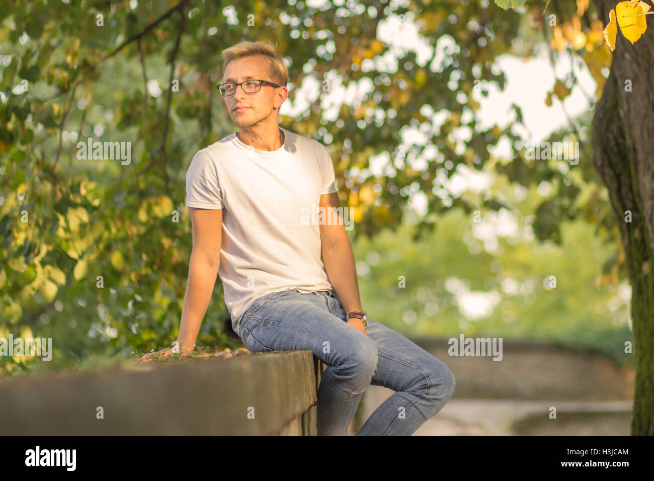Día soleado de verano al aire libre un joven sentado ropa casual camiseta blanca jeans Foto de stock
