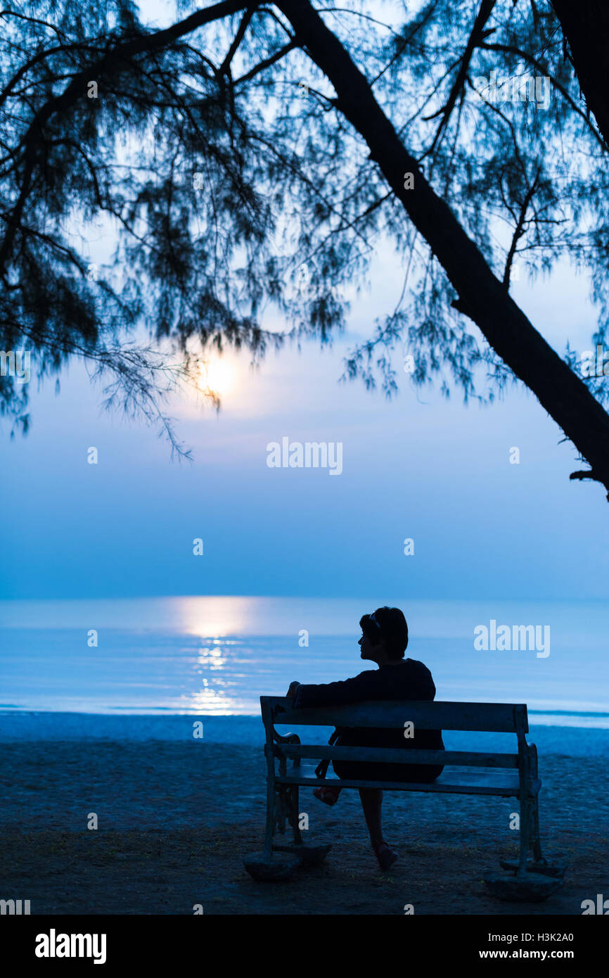 Soledad, silueta mujer viajero sentado solo con su bolsa en la banqueta retro durante la noche en la playa. Foto de stock