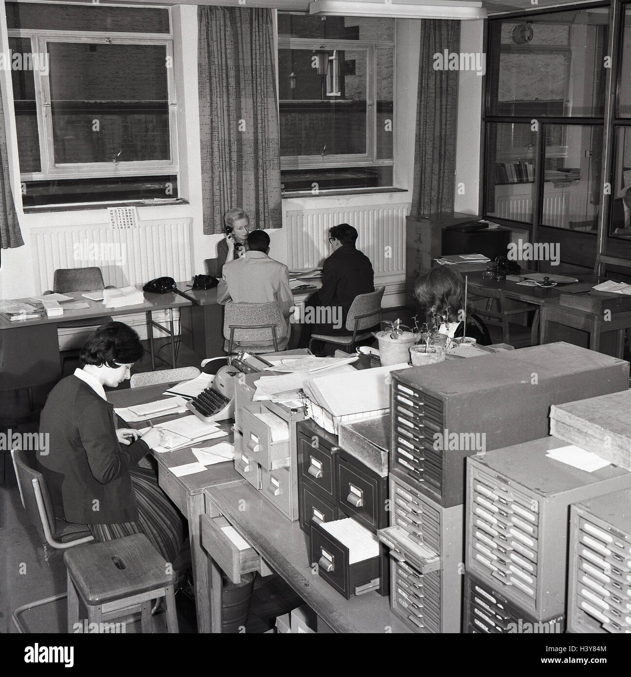 1960, históricos, consejo local, departamento de administración, secetary en escritorio con máquina de escribir junto a varias filas de cajas de índice de tarjeta. Foto de stock