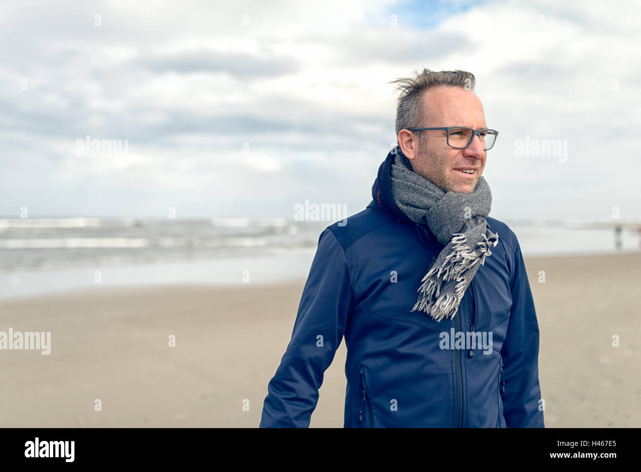 Irritado hombre de mediana edad con gafas y una bufanda de lana tejida de pie en una desierta playa de otoño en un día nublado con c Foto de stock