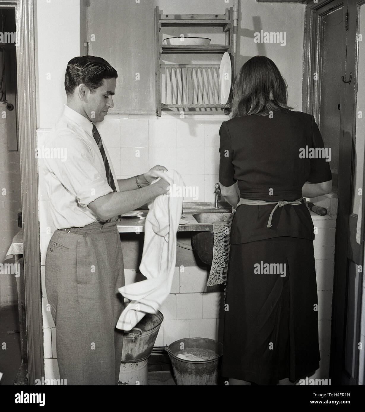 1950, en este período histórico, vemos un hombre con un paño húmedo secado placas, como una dama limpia los platos en el fregadero en el lavadero, Londres, Inglaterra. Foto de stock
